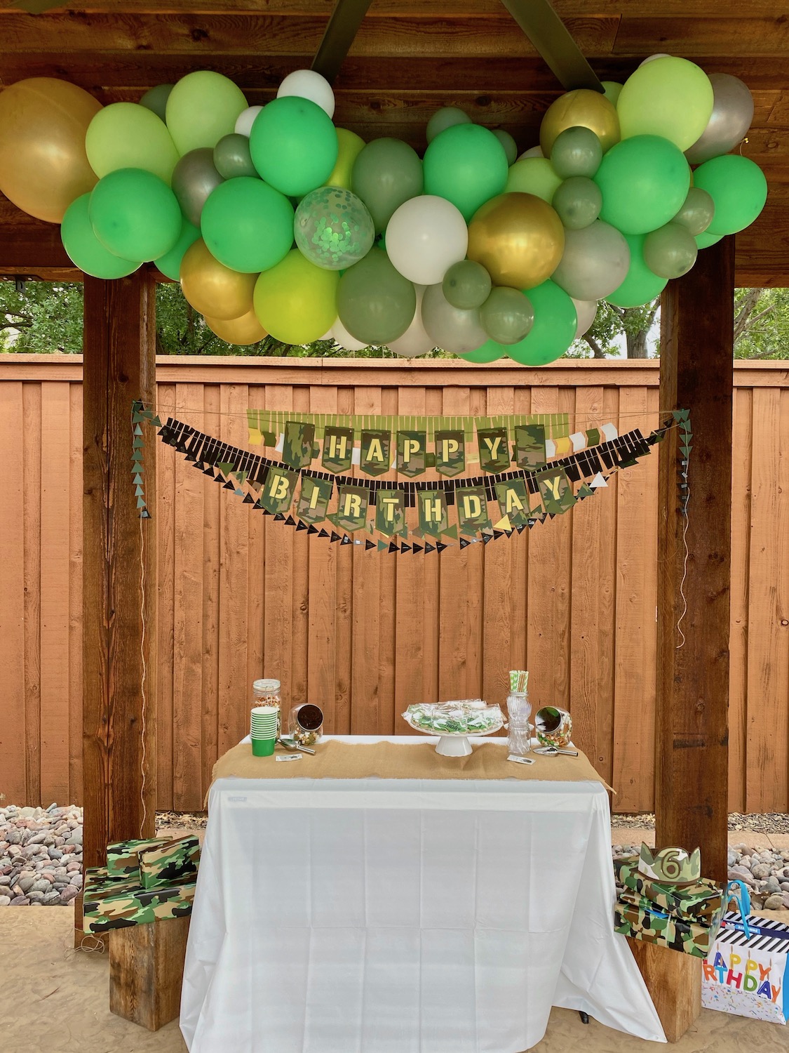 A table with a white tablecloth and burlap table runner. A green balloon garland hangs above the table with a camouflaged themed happy birthday banner below it. On the table are birthday party snacks including vanilla cupcakes, carrot cake with cream cheese frosting and camouflage themed sugar cookies.