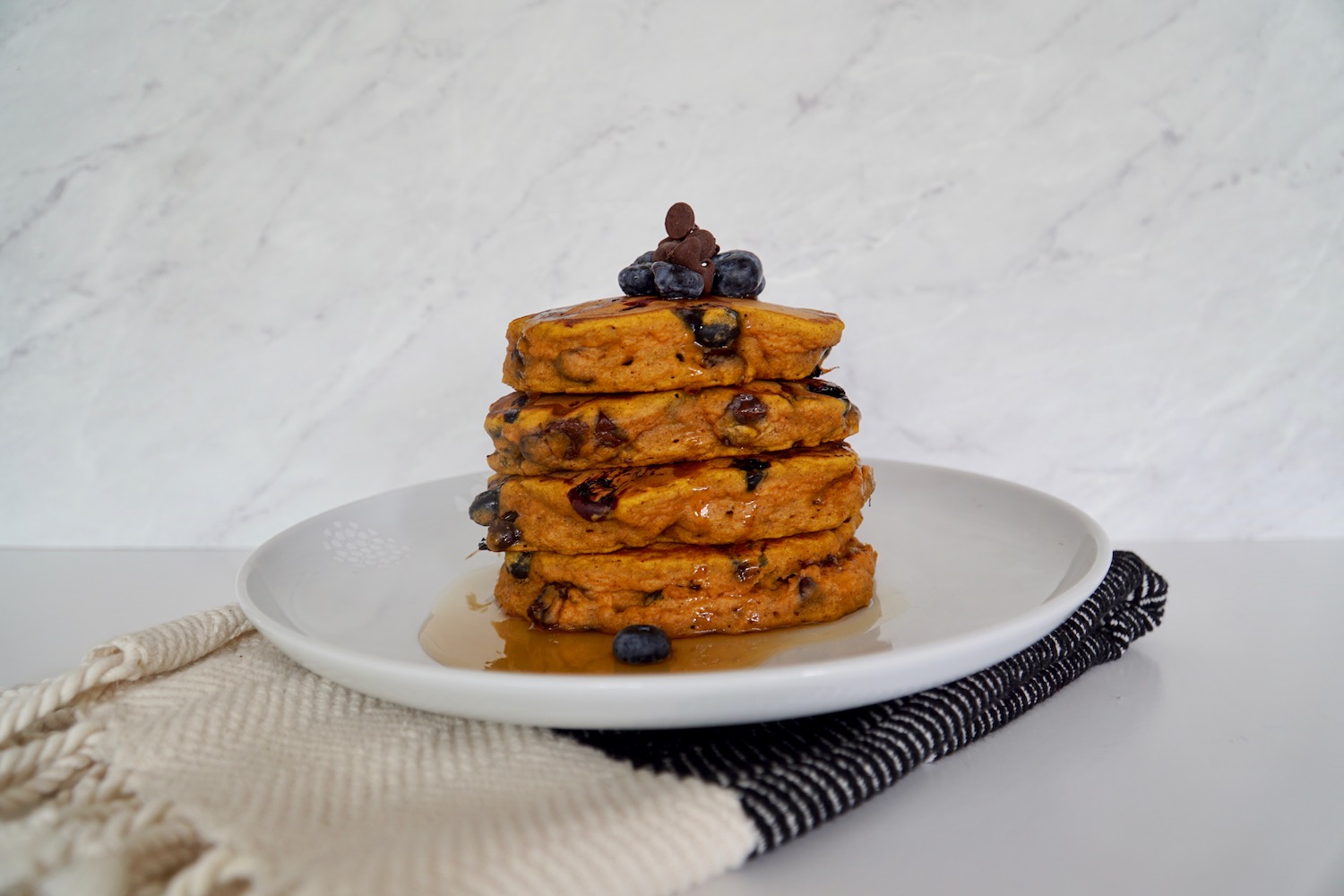 Stack of 4 Pumpkin-honey pancakes with syrup, blueberries and chocolate chips sitting on a white plate on top of a kitchen towel.